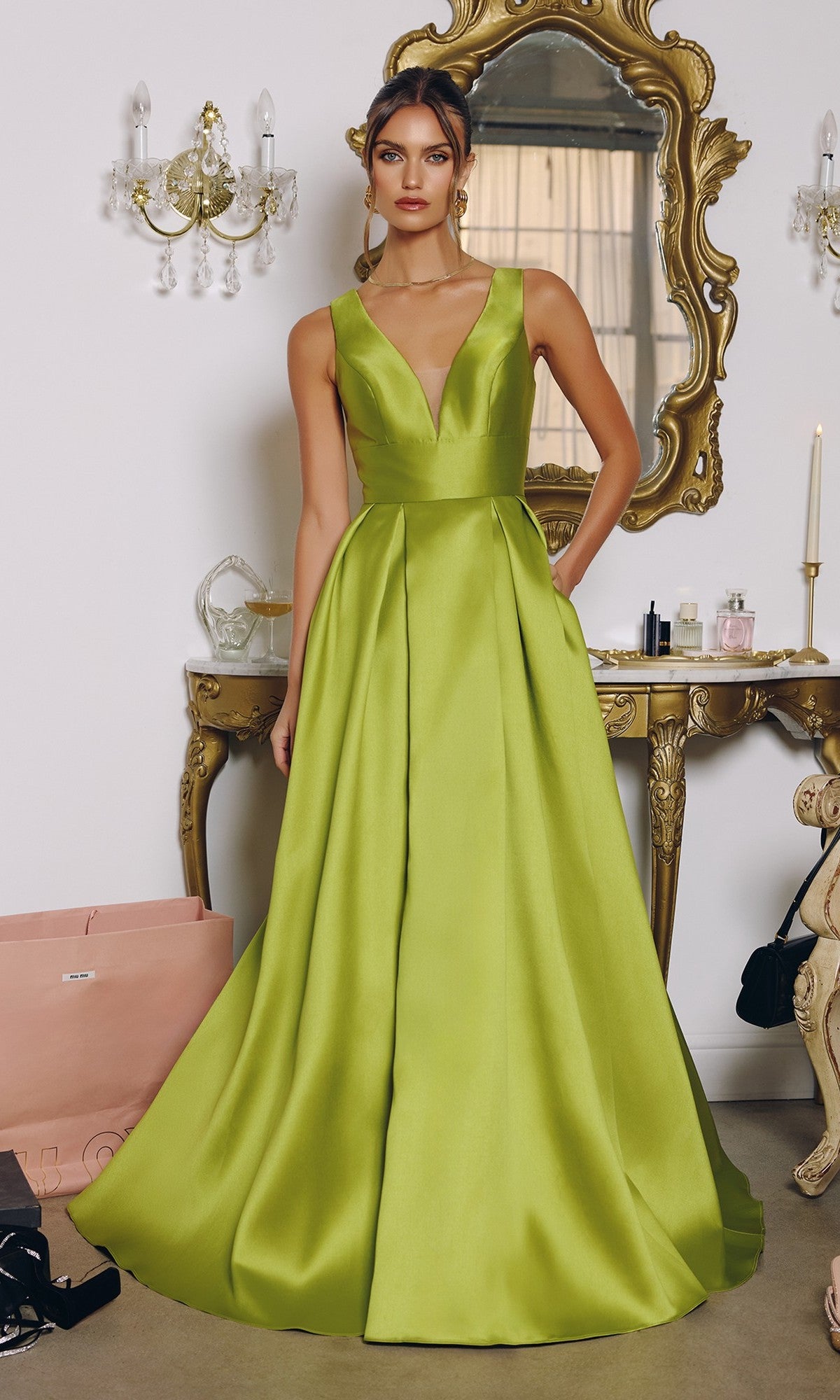 A woman stands indoors wearing the Amoris T814 long prom dress in Greenery, a sleeveless satin A-line gown with a deep V neckline, in front of a gold-framed mirror and ornate vanity table.
